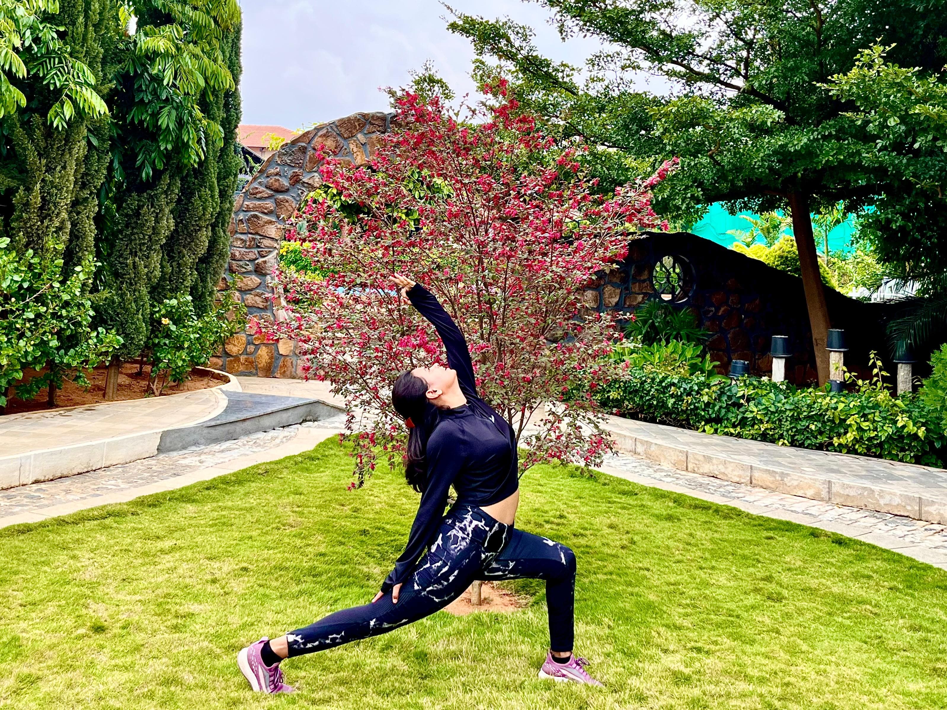 Shiya Shahi in a crescent lunge with one arm raised in a garden